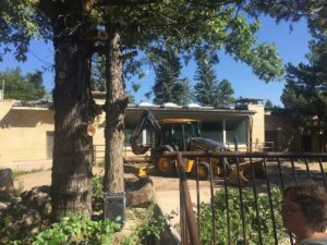 Yellow backhoe by brick building and trees